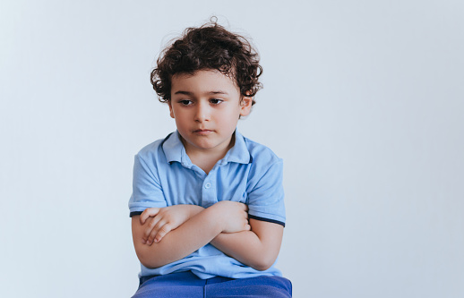 Confused curly caucasian boy in blue polo sitting hands folded on chest looks down against white wall. Handsome sad toddler upset isolated at studio. Frustration, childhood.