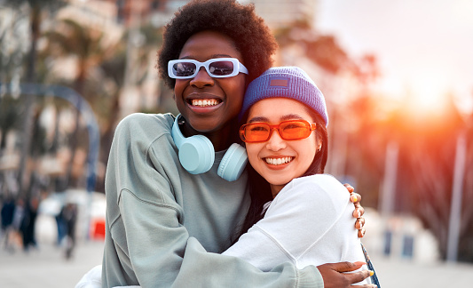 African American and Asian young cute women girlfriends hugging and having fun outdoors listening to music in headphones.