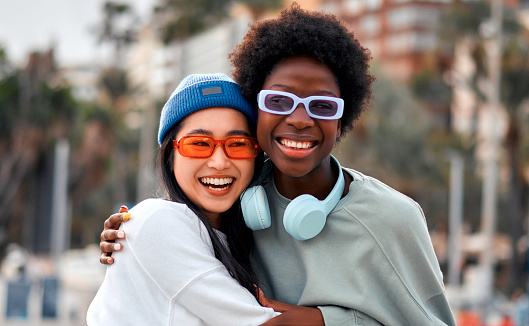 African American and Asian young cute women girlfriends hugging and having fun outdoors listening to music in headphones.