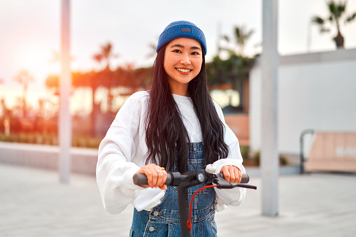 Pretty Asian young woman in a hat riding a scooter on the streets of the city. Active recreation and sports.