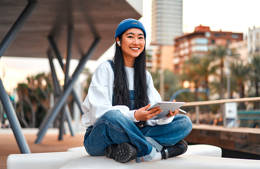Cute Asian young woman in a hat with a tablet on the street. Drawing on a graphics tablet. Freelancing and earning online.
