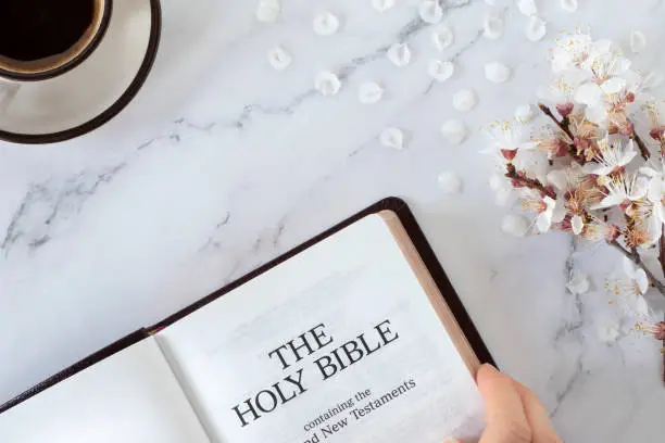 Hand holding open holy bible book, cup of coffee, and spring branch with white flowers on marble background. Top table view. Symbol of Christian new life and growth.