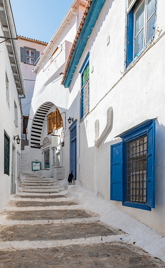 Narrow street with steps between white buildings in Hydra town, Greece. Window with blue shutters. Diminishing perspective.