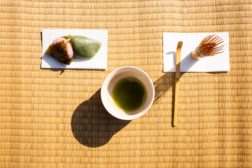 Tea utensils lined up on tatami mat goza.\nChashaku (tea scoop), chasen (tea whisk), kaishi (tea paper), tea bowl, and wagashi (Japanese sweets).\nCasual style Nodate (outdoor tea ceremony).\nDomyoji wagashi (Japanese sweets) and yomogi mochi (mugwort rice cake).\nThe tea bowl contains matcha.\nA traditional Japanese event.\nTaken outdoors.\nTaken from above with a bird's eye view.
