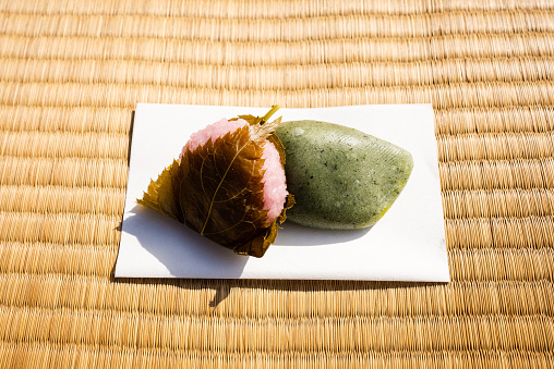 Domyoji wagashi (Japanese sweets) and yomogi mochi (mugwort rice cake) are served on a piece of kaishi paper.\nA scene of Nodate (outdoor tea ceremony) with cherry blossoms.\nJapanese tea ceremony outdoors in a casual setting.\nCherry blossom viewing is a seasonal event in Japan.