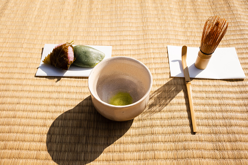 Tea utensils lined up on tatami mat goza.\nChashaku (tea scoop), chasen (tea whisk), kaishi (tea paper), tea bowl, and wagashi (Japanese sweets).\nCasual style Nodate (outdoor tea ceremony).\nDomyoji wagashi (Japanese sweets) and yomogi mochi (mugwort rice cake).\nThe tea bowl contains matcha powder.\nA traditional Japanese event.\nTaken outdoors.