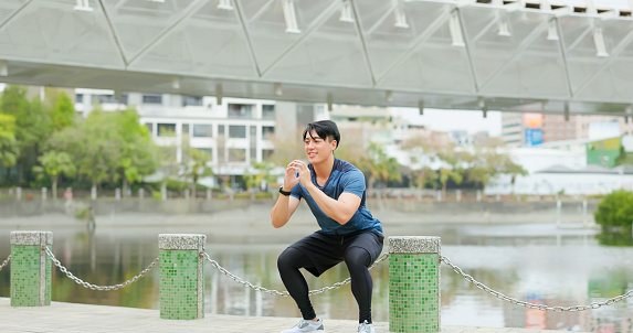 asian smiling young man is doing squat exercise outdoor in city