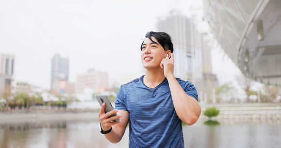 asian male runner wearing earbuds and using mobile phone to listen music is ready to jog in city