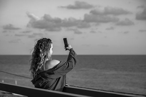 a beautiful blonde woman with long wavy hair stands on a bridge and shoots a sunset at the sea on her phone, black and white photo
