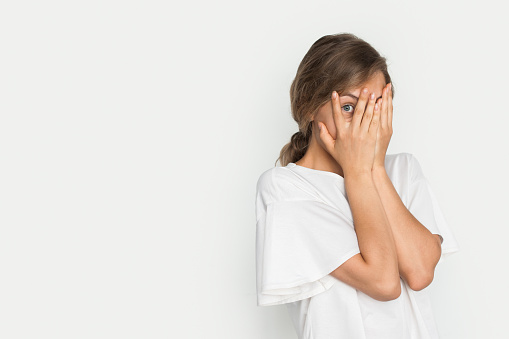 Young pretty blonde woman peeking through her fingers isolated over white background with copy space. Shy teen girl covering face with hands.