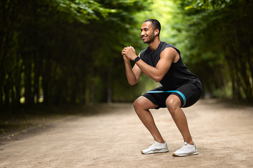 Motivated handsome muscular millennial black man exercising in public park, using elastic fitness band, enjoying outdoor workout, panorama with copy space. Sports lifestyle and millennials
