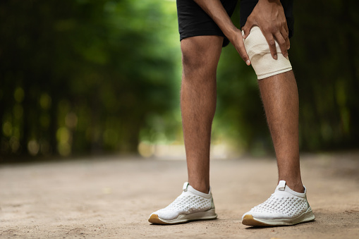 Knee injury and workout concept. Unrecognizable sportsman wrapping injured knee with elastic bandage before training, exercising in public park, panorama with copy space, cropped