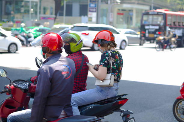 Young thai woman with protective face mask is using mobile phone while sitting in back of motorcycle taxi which is waiting at junction of Phetchaburi Road in Bangkok. In background is traffic