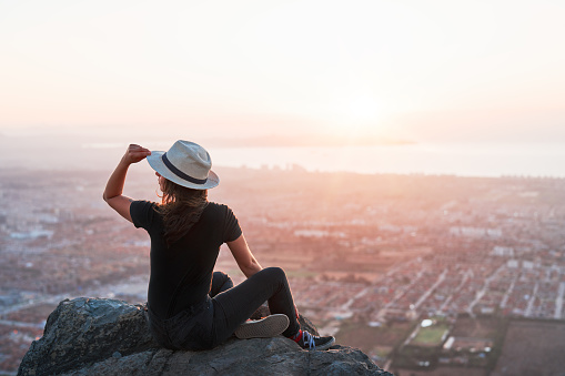 rear view portrait young woman sitting on a rock at the viewpoint on top of a hill overlooking the city and or ocean on the horizon
