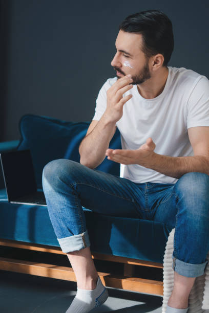 ritratto di giovane bell'uomo che mette la crema sul viso per mantenere la sua pelle in buona forma, seduto sul divano nel soggiorno. concetto di cura del corpo. - men smooth the human body moisturizer foto e immagini stock