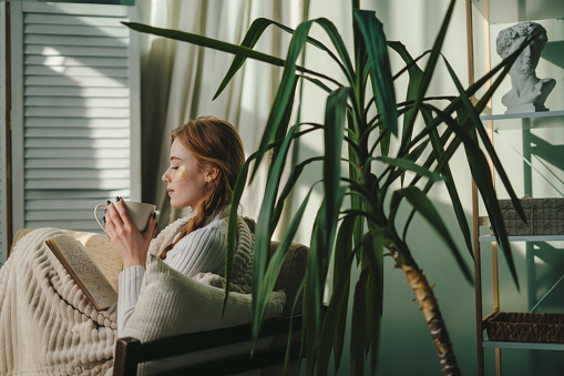 Sideways young woman drinking coffee and reading book sitting on sofa while staying at home, spending her spare time in living room, indoor. People, season and leisure concept