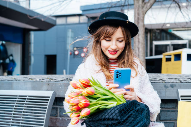 istock Stylish smiling young woman using her phone for messaging, communication, content creating for social media sitting bench outdoors. Girl holding tulips bouquet. Springtime street fashion. Influencer. 1474877646
