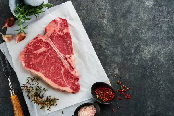Photo of Thick Raw T-Bone Steak. Dry-aged Raw T-bone or porterhouse beef meat Steak on parchment paperwith herbs and salt on dark background. Top view and copy space.