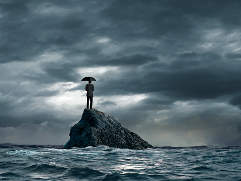 A man holding an umbrella stands on a tiny rocky island as he looks out toward the distant horizon as rough water start to churn below him. A stormy sky conveys a sense of foreboding.