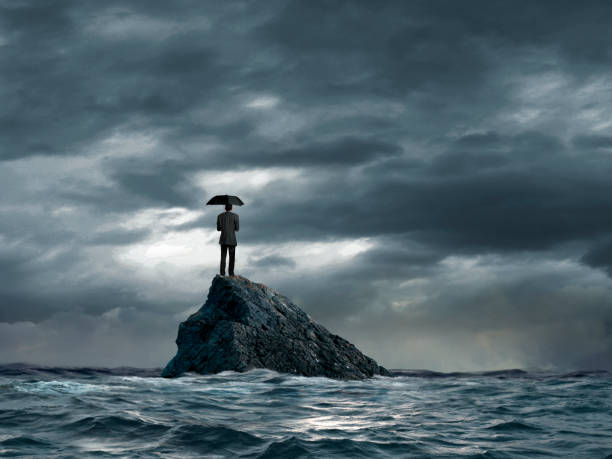 Man Stranded In On Tiny Rock Holding Umbrella A man holding an umbrella stands on a tiny rocky island as he looks out toward the distant horizon as rough water start to churn below him. A stormy sky conveys a sense of foreboding. calm before the storm stock pictures, royalty-free photos & images