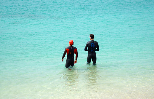 Playa Blanca, Lanzarote, Spain - March 18. 2022: Two male swimmers in wetsuits facing out to sea in bright sunshine.