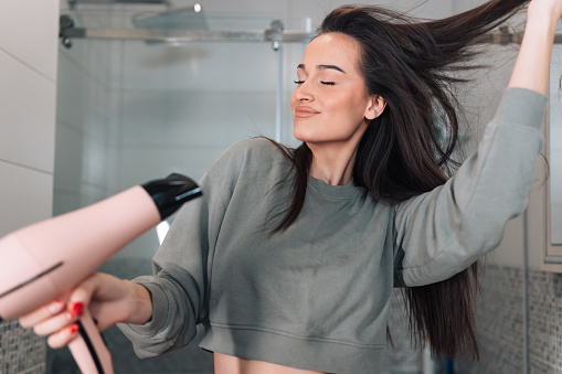 Beautiful young woman in a good mood blow drying hair in the bathroom