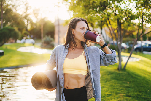 Beautiful Caucasian woman holding a yoga mat in the park drinking a cup of tea or coffee and looking away. Healthy lifestyle, wellness. Healthy lifestyle, sport. Training workout. Fitness woman.
