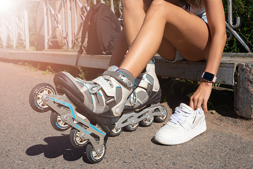 Close up girl putting on inline skates rollers preparing in public park in summer. Teenager leisure outdoor sport activity game.