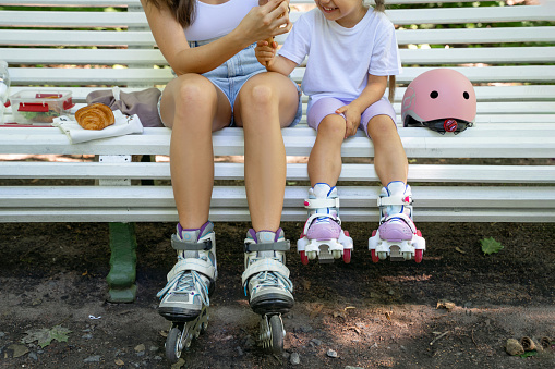 Mother and daughter having snack on bench after skating inline roller in public park together. Family leisure sport activity weekend. Copy negative space