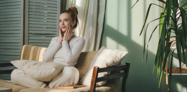 Satisfied young woman applying moisturizing cream on face, posing on couch against cozy interior. Natural beauty. Home comfort. Healthy body care. Skin care.