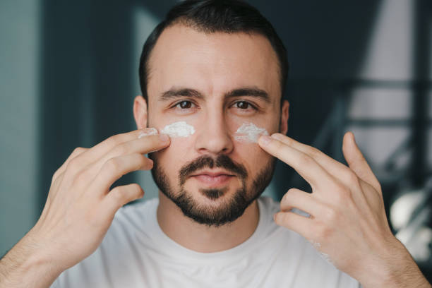 ritratto ravvicinato di un uomo caucasico soddisfatto che applica la crema di routine per la cura della pelle sul viso, guardando la fotocamera. bellezza del viso. trattamento viso. stile di vita felice. cura sana del corpo. assistenza sanitaria, trattamen - men smooth the human body moisturizer foto e immagini stock