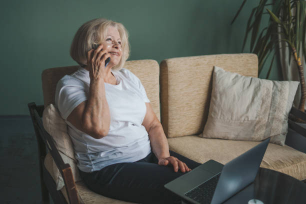 istock Active smiling old woman talking on mobile phone while using laptop in living room at home. Woman surfing internet. Online working. 1474885317