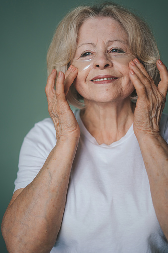 Smiling old caucasian woman with natural beauty applying patches under her eyes. Face skincare in older age. Beauty face. Anti-aging skincare. Natural beauty.