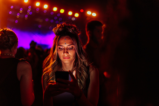 A young Caucasian woman is using her smartphone at a concert, reading text messages with a serious face illuminated by her device.