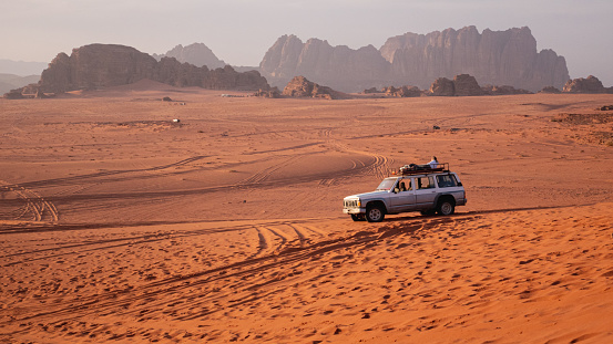 A 4x4 vehicle standing in the Wadi Rum desert with man sitting on top of it, in the background tall rock formations are seen.