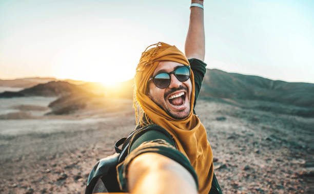 Handsome man taking selfie pic with smart mobile phone outside at golden hour time - Traveler guy with backpack enjoying day out on summer vacation - Happy tourist having fun at summertime holiday stock photo
