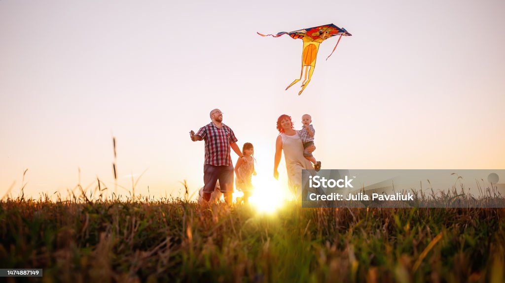 Panoramic diversity view of family running at sunset with kite in the sky. View from below Panoramic diversity view of family running at sunset with kite in the sky. View from below through the ears. Silhouette. The father holds toy in hands, the mother hugs little son, the daughter has fun Kite - Toy Stock Photo