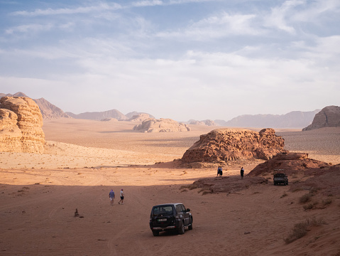 A group of parked 4x4s parked in Wadi Rum in the shade waiting for tourists to come back, in the background rock formations in the sun are visible.