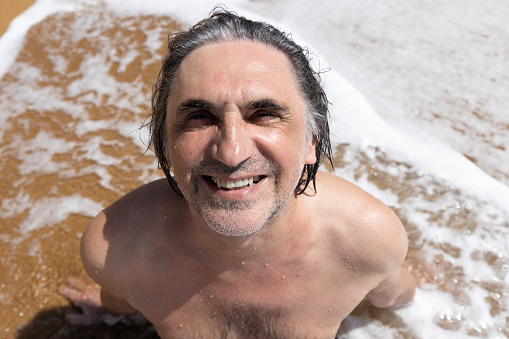 Adult man sitting on yellow sand in a foam wave, Greece, Crete, Chania. High quality photo