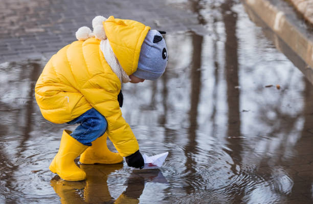 istock A small child in yellow rubber boots and a jacket runs through puddles, has fun, plays and launches paper boats. Spring break photo. It's springtime. 1474895113
