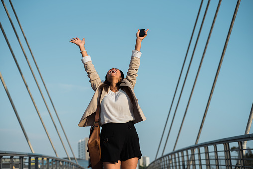 Latina woman raises her arms in triumph after receiving good news on her cell phone. Copy space