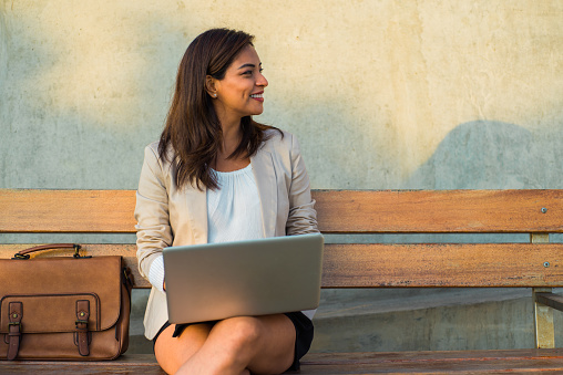 A Latina woman sitting on a bench with her computer looks smiling at the copy space.