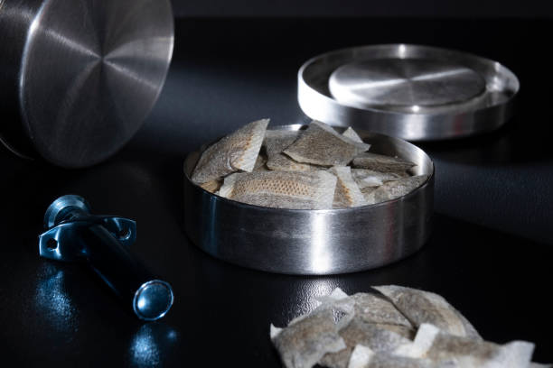 Closeup of a metallic Swedish snus can with white portion snus pouches. A blue loose snus portioner in the foreground stock photo