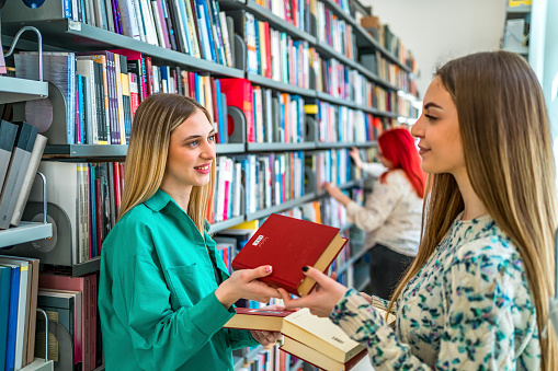 Cheerful female students exchanging books and talking in the college library