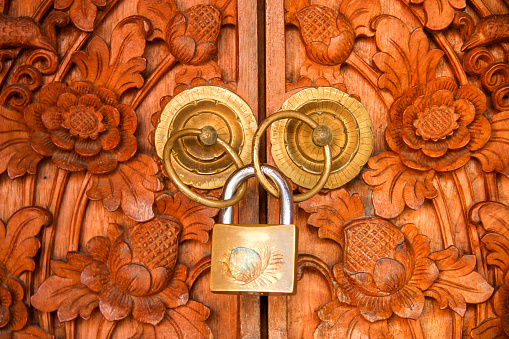 Horizontal closeup photo of intricately carved floral pattern wooden doors, shut with a brass padlock.