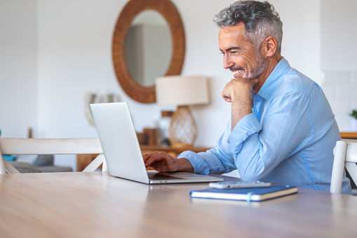 Mature businessman working on a laptop at home. He is happy and smiling and casually dressed.