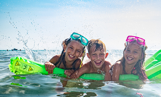 A boy and two girls bathe together in the sea on a mattress, sunbathe, laugh together, and enjoy themselves.