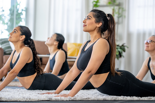 A small group of women lay out on the floor in a living room as they practice Yoga together.  They are each dressed comfortably in athletic wear as they hold their pose.