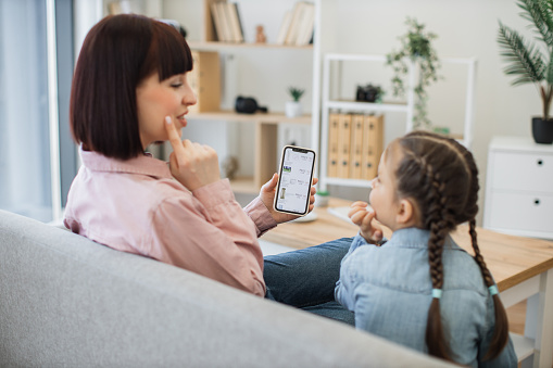 Beautiful young mother teaching her little daughter main rules of choosing cosmetics while browsing online boutique during sale season. Happy family having fun while doing home shopping on smartphone.