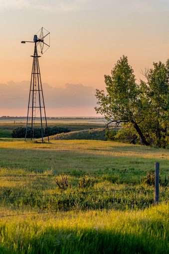 Sunset over an old windmill in a pasture at sunset with trees and a fence line in the foreground